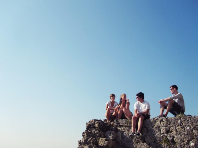 Vier junge Menschen sitzen auf einem Felsvorsprung, im Hintergrund hellblauer Himmel.